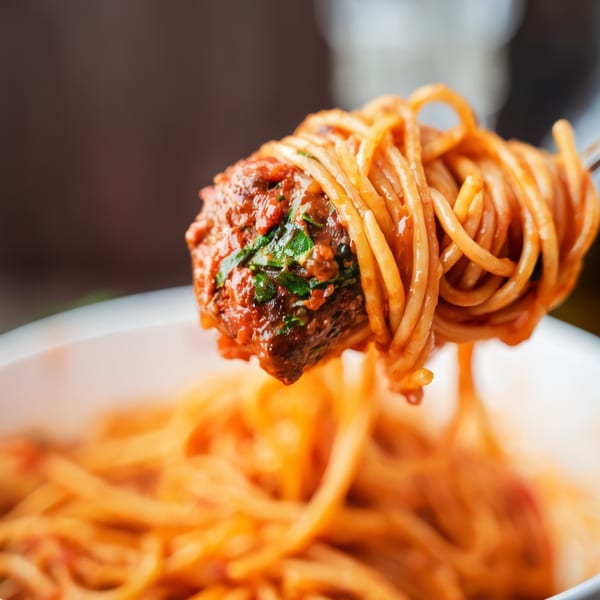 A fork lifts a large meatball and spaghetti covered in red sauce from a white bowl.